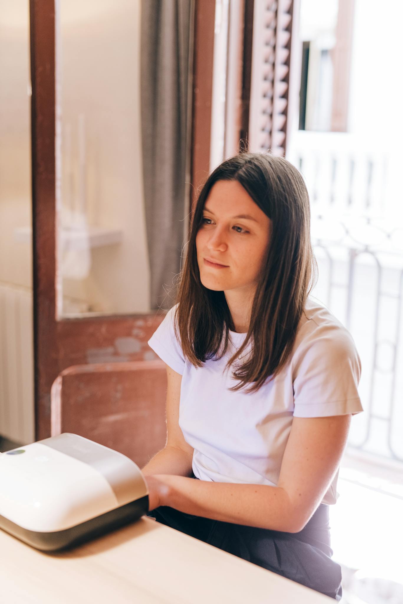 A woman with brunette hair uses a nail dryer indoors during the day.