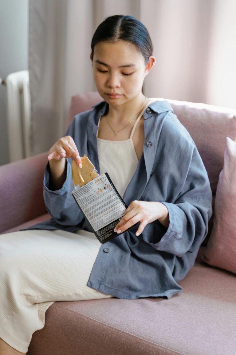 Asian woman sitting on a couch unpacking a skincare product with focus and care.